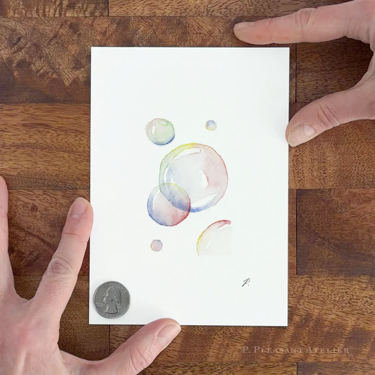 A hand-painted watercolor art print of translucent bubbles on white paper, held down by two hands over a wooden surface with a coin for scale.