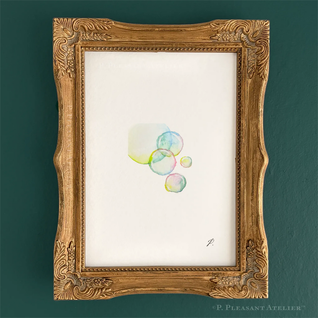 A framed art print featuring a delicate watercolor illustration of pastel-colored bubbles on textured white paper, housed in an ornate gold frame against a dark teal wall.