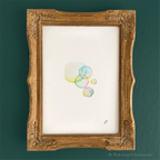A framed art print featuring a delicate watercolor illustration of pastel-colored bubbles on textured white paper, housed in an ornate gold frame against a dark teal wall.