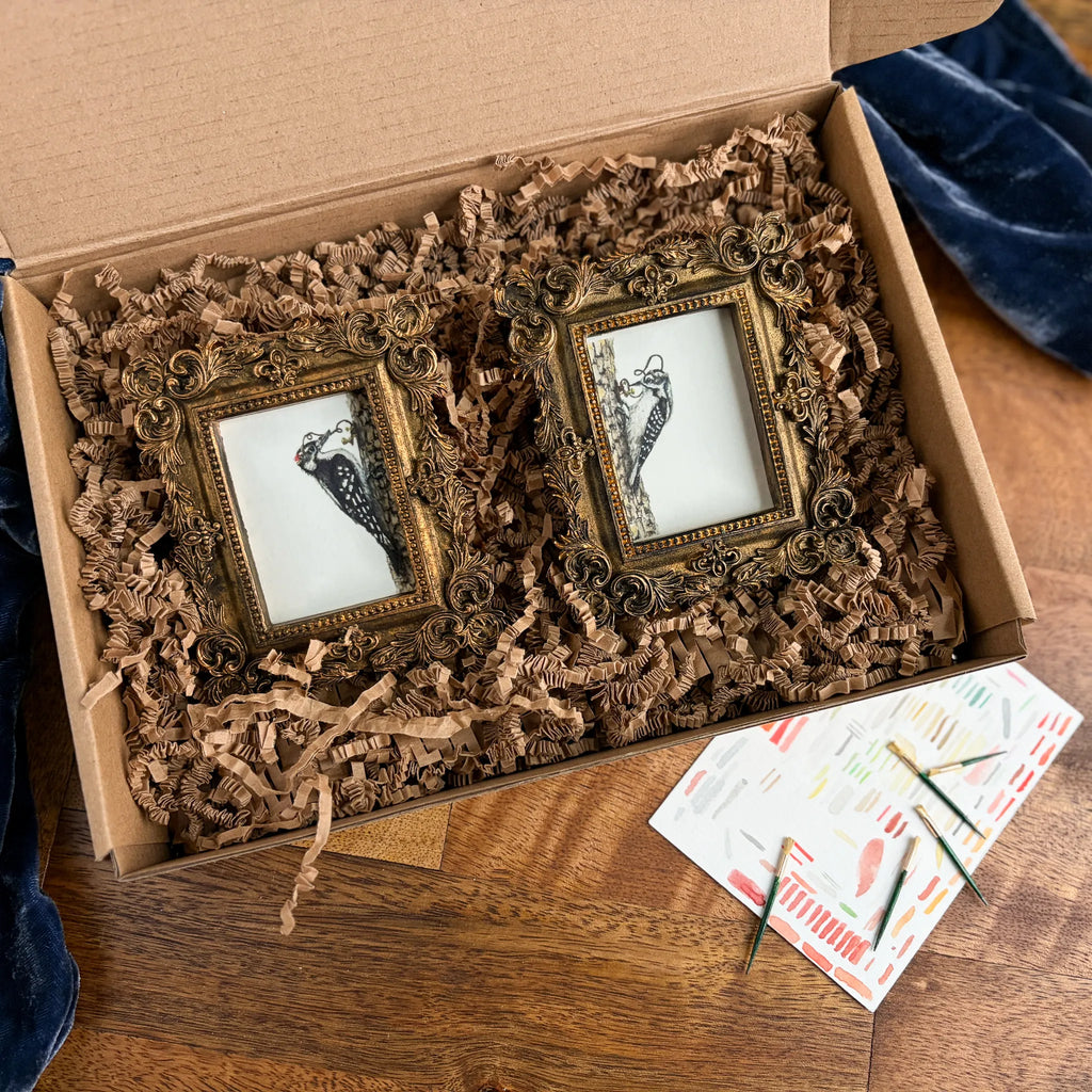 An open cardboard gift box filled with brown paper shred contains two small ornate gold picture frames with black-and-white bird illustrations. A printed card with small green and gold items lies on the wooden table beside the box.