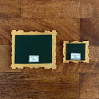 Two decorative square frames with scalloped gold borders and dark green textured centers, displayed on a brown wood plank background. Each frame has a small white label reading 'P. Pleasant Atelier.'