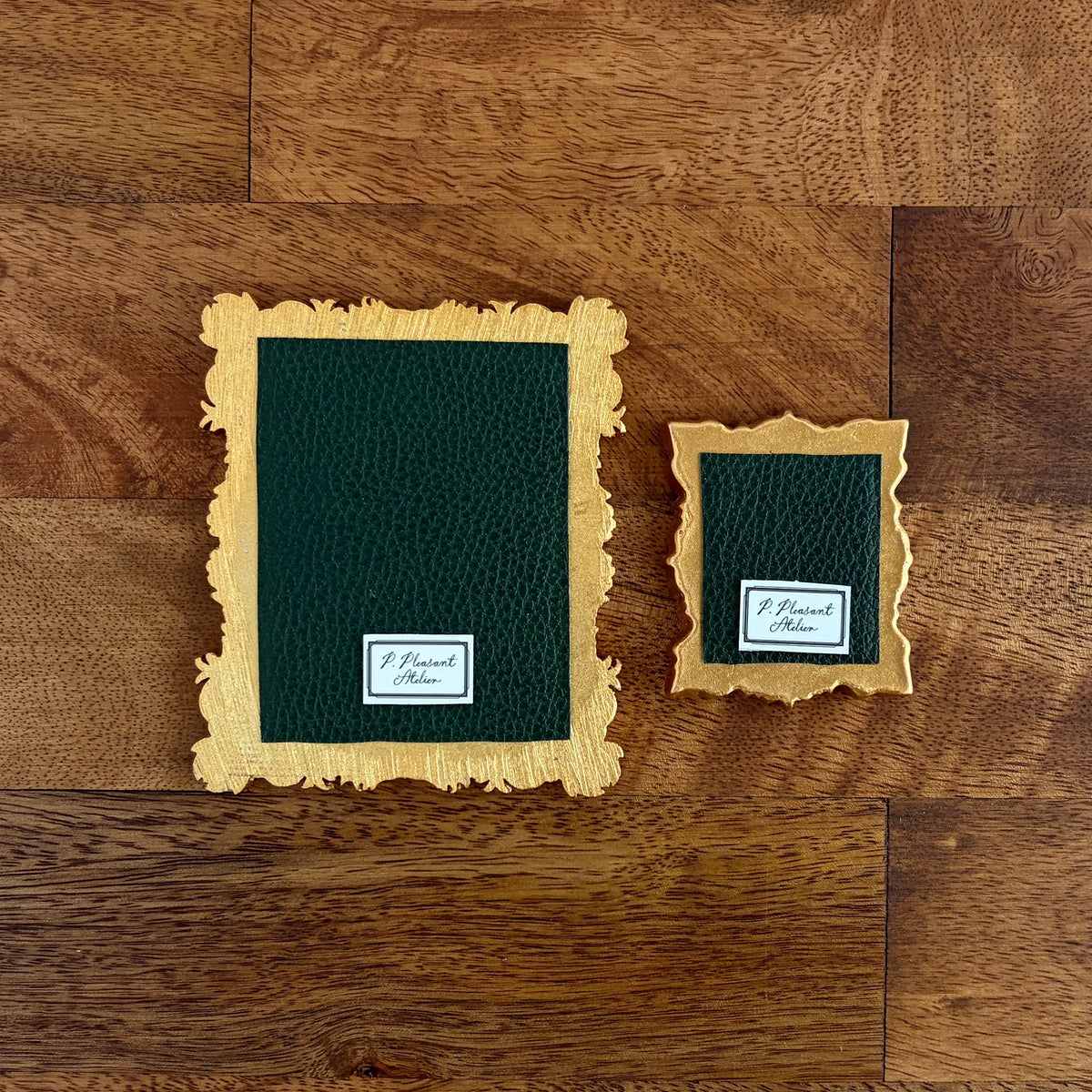 Two decorative picture frames with textured dark green centers and scalloped gold edges, displayed on a brown wood background. One frame is large and one is small, both featuring a white label at the bottom that reads 'P. Pleasant Atelier'.