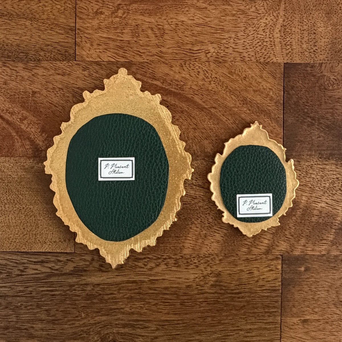 Two oval ornate gold frames and dark green textured centers, displayed on a brown wood plank floor. Each piece has a small white label reading 'P. Pleasant Atelier.'