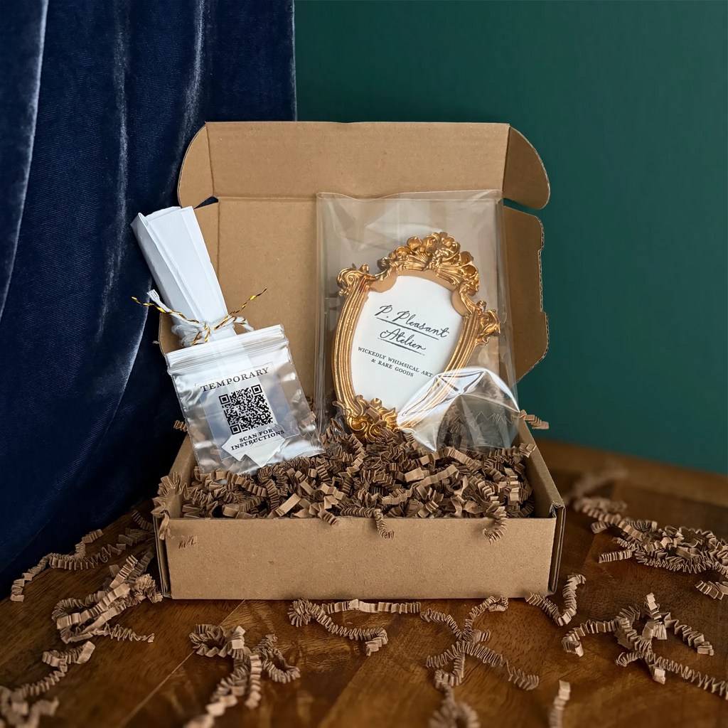 A curated gift box set in a brown cardboard box with shredded paper filler, featuring a small ornate gold frame with a wedding invitation insert, a QR code favor bag, and a wrapped favor, set against a dark teal curtain and wooden table.