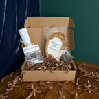 A curated gift box set in a brown cardboard box with shredded paper filler, featuring a small ornate gold frame with a wedding invitation insert, a QR code favor bag, and a wrapped favor, set against a dark teal curtain and wooden table.