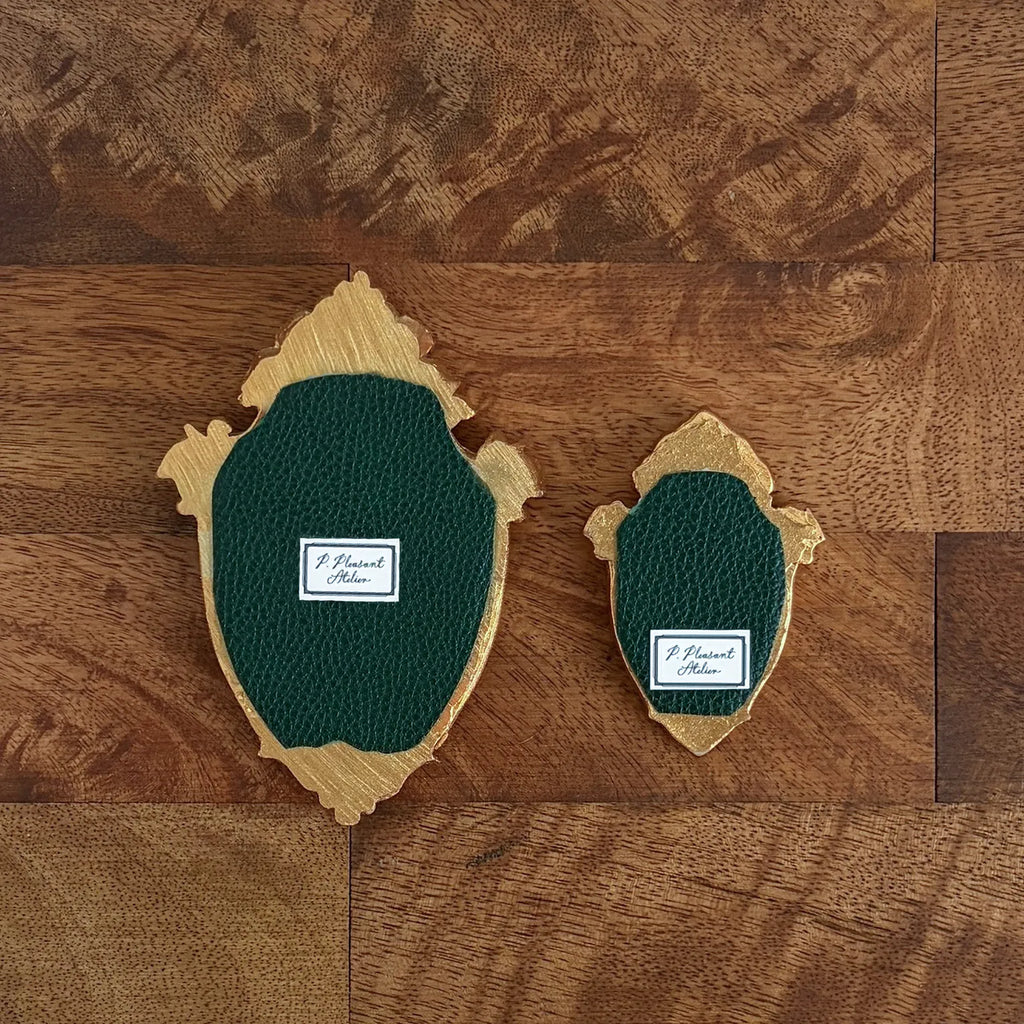 Two decorative shield-shaped wall pieces with dark green textured center panels and natural wood edges, displayed on a brown wood plank background. Each piece has a small white label reading 'J.