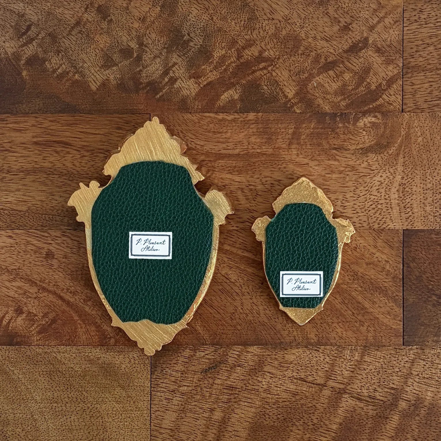 Two decorative shield-shaped wall pieces with dark green textured center panels and natural wood edges, displayed on a brown wood plank background. Each piece has a small white label reading 'J.