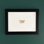 Art print in a dark wooden frame on a teal wall, with a small cup shaped emblem near the bottom