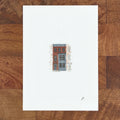 Art fairy magic illustration of a small red and blue building on a white paper sheet on a wooden table